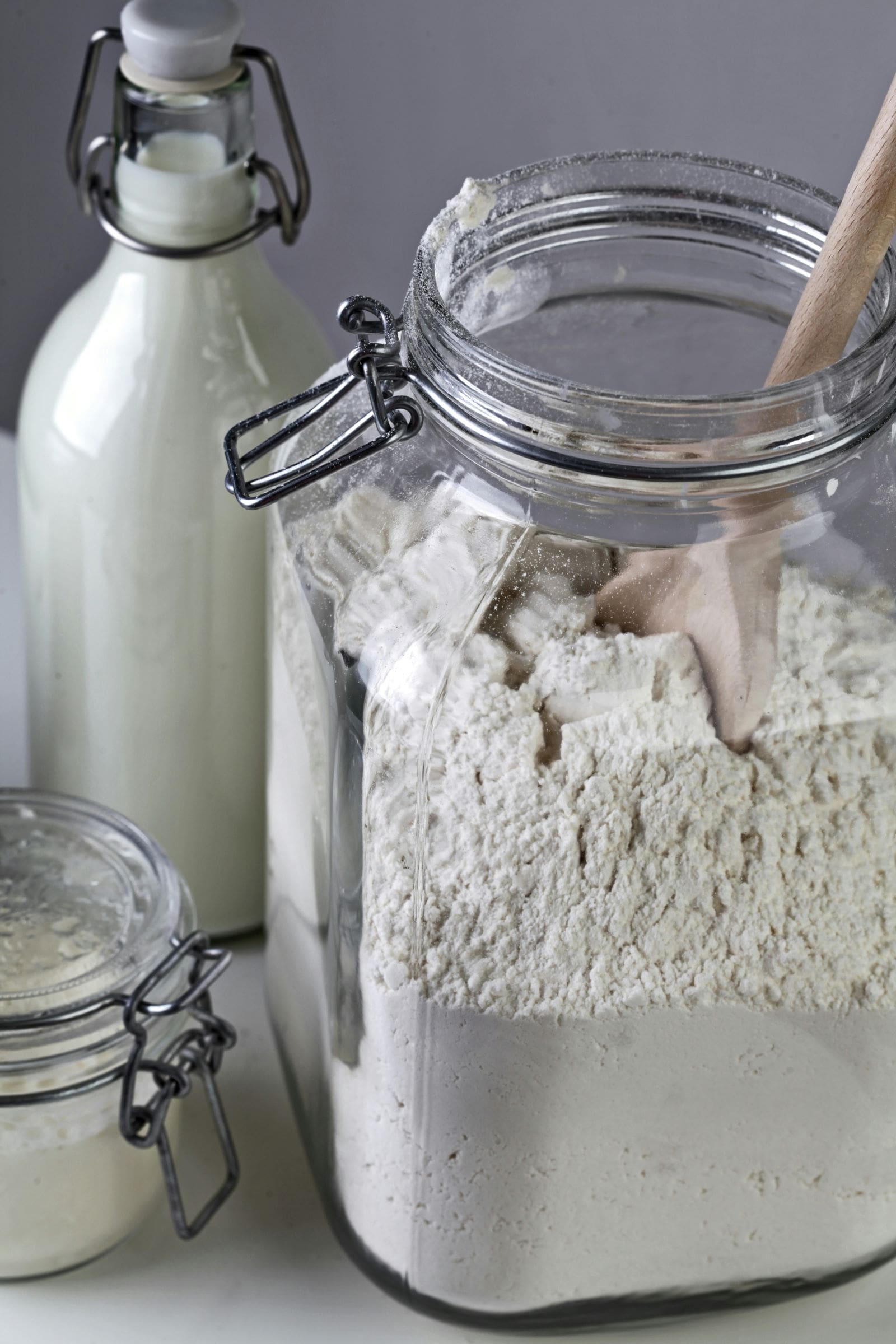 A jar of flour, a jar of milk, and a wooden spoon arranged on a countertop, ready for baking preparations.