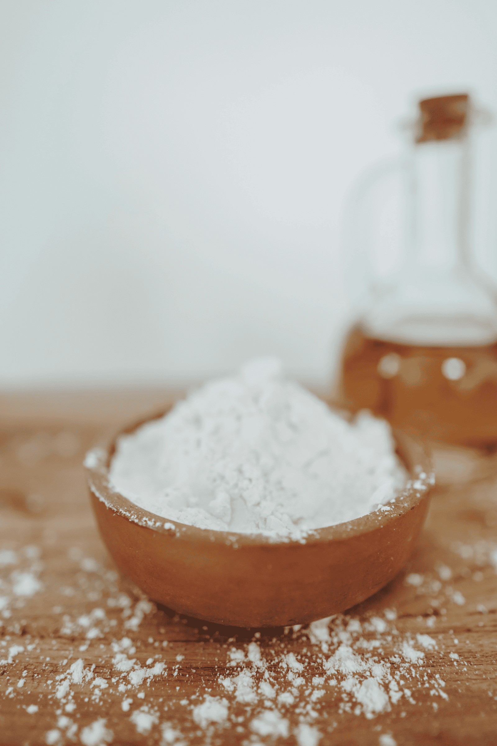 A wooden bowl filled with white flour sits on a wooden surface, with scattered flour around it. A glass carafe of oil is blurred in the background.