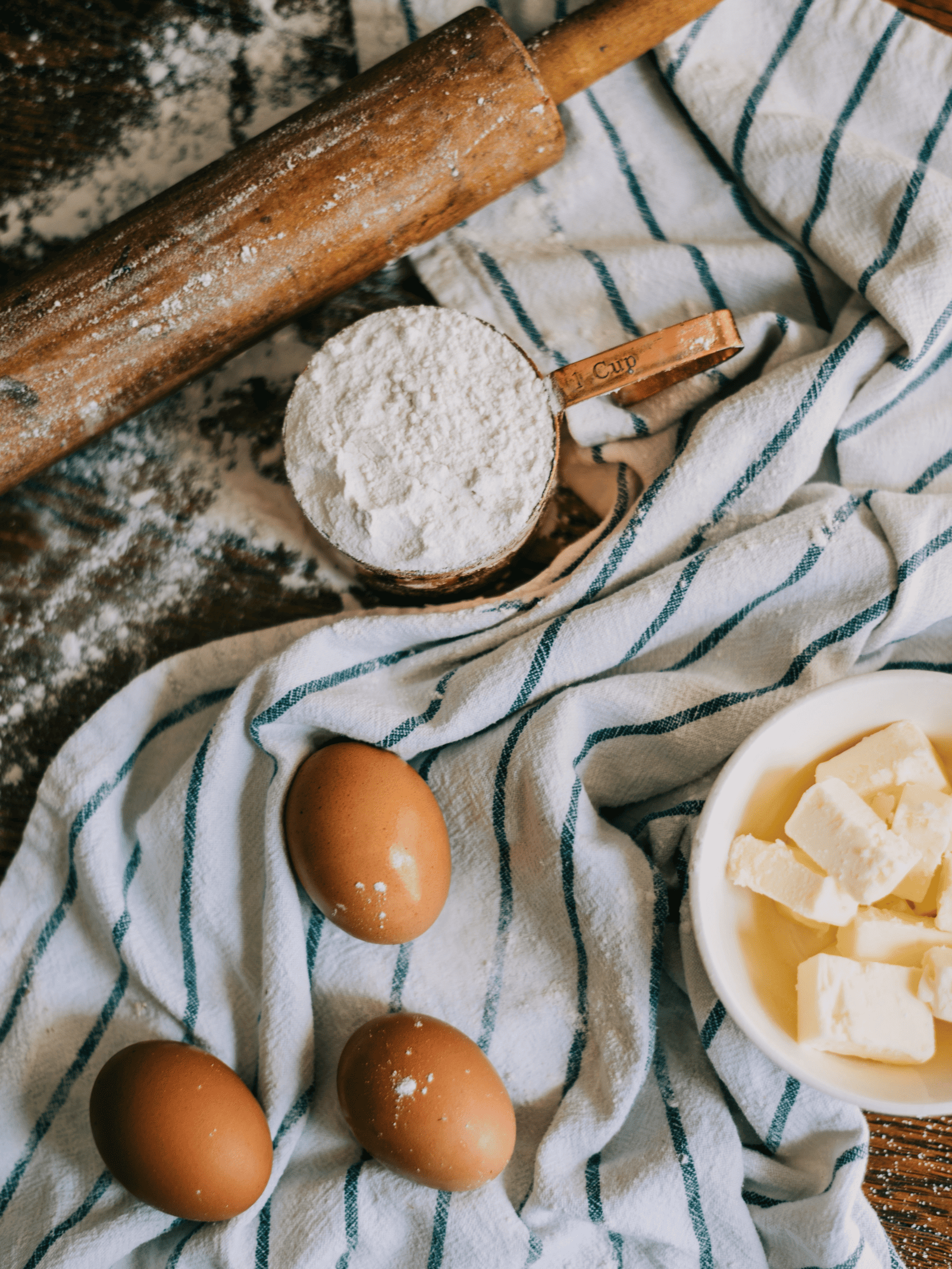 A cozy baking scene with three eggs, a dish of butter cubes, a copper cup of flour, and a wooden rolling pin on a striped cloth, evoking warmth.