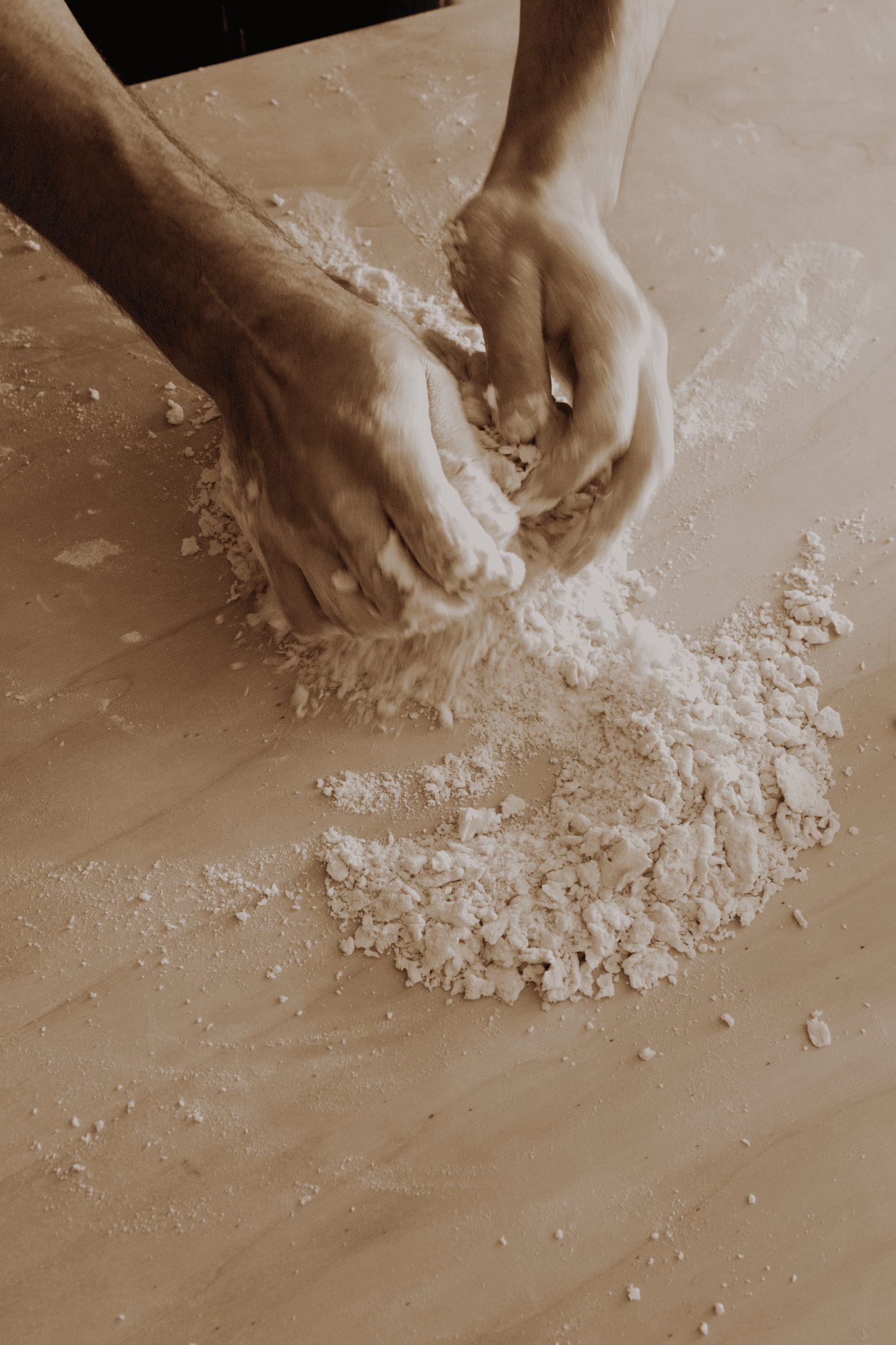 A person’s hands kneading and mixing dough on a wooden surface dusted with flour.