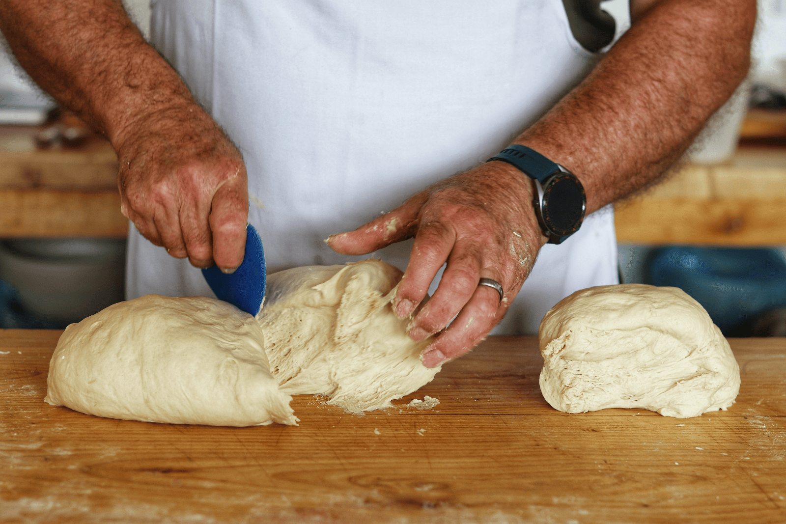 Hands of a person in a white apron dividing dough on a wooden surface with a blue dough scraper. The scene conveys focus and craftsmanship