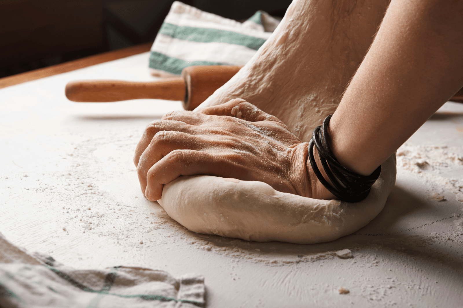 Close-up of a hand kneading soft dough on a floured surface, with a wooden rolling pin and striped kitchen towel in the background, conveying a sense of home baking