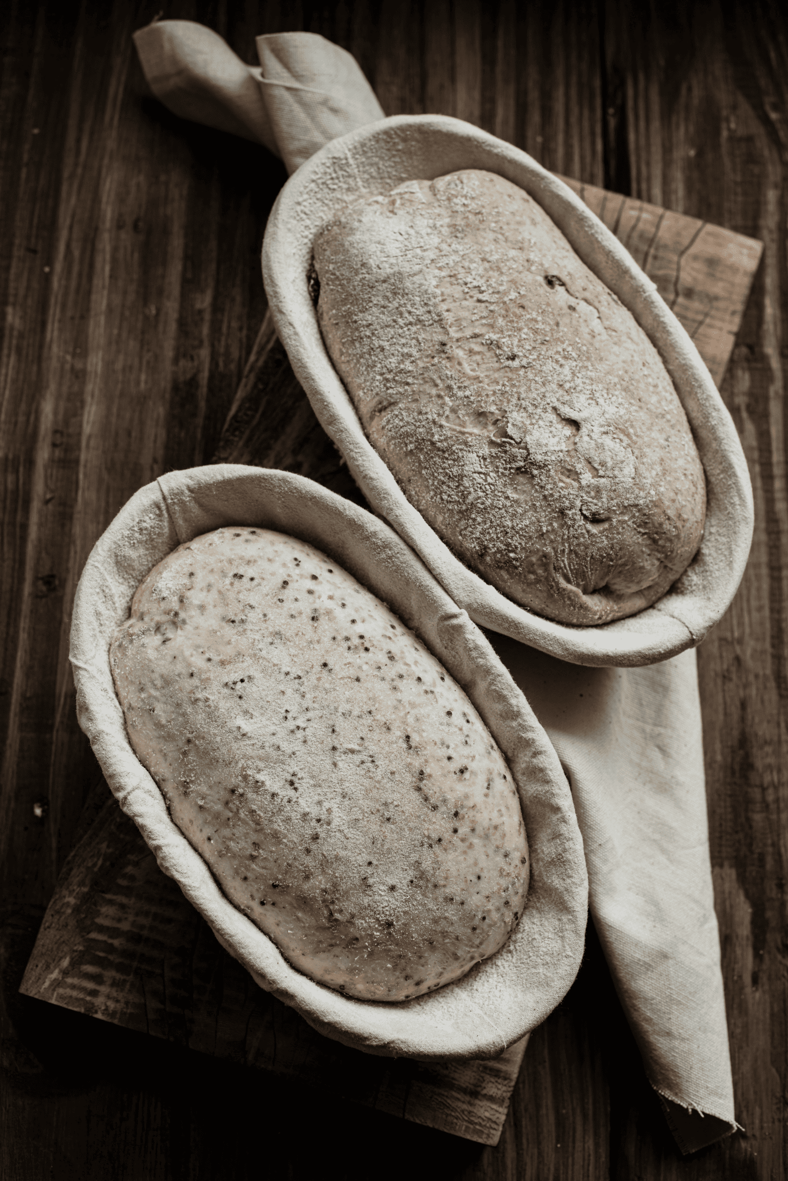 Two oval-shaped loaves of bread dough rest in cloth-lined proofing baskets on a wooden surface, ready for baking—a perfect scene unless you're left wondering, why is my bread not rising?