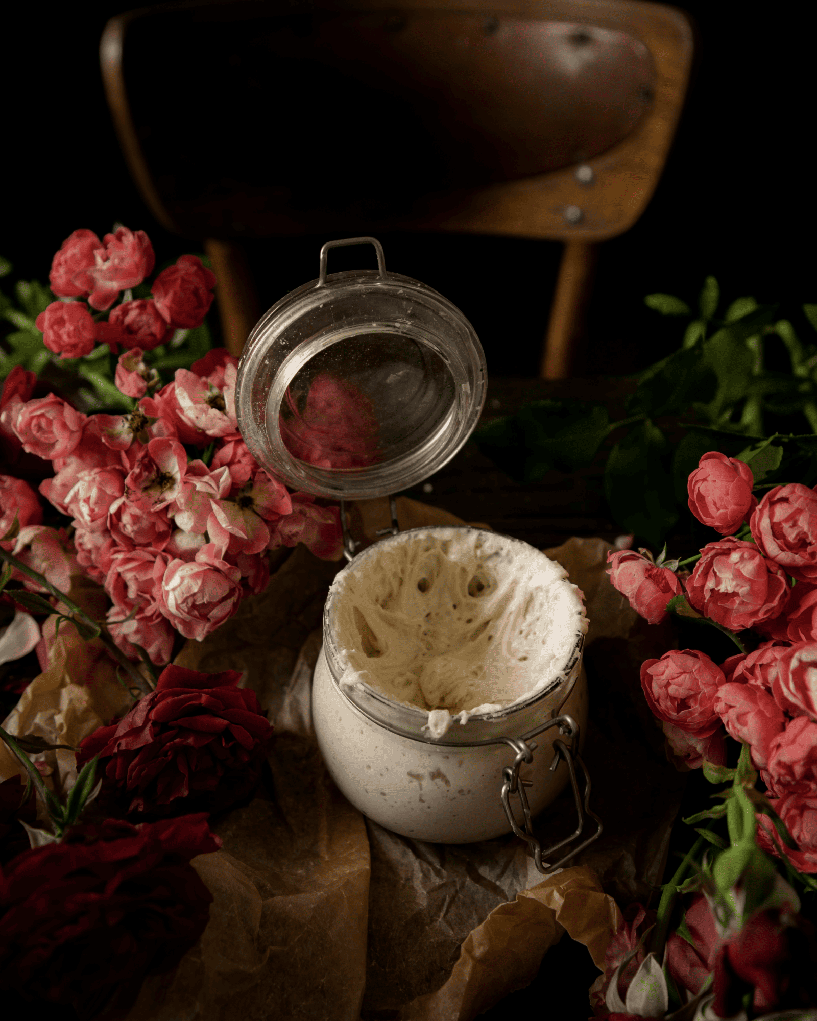 An open glass jar of yeasty dough sits among pink and red roses on brown parchment paper, evoking a cozy and romantic atmosphere
