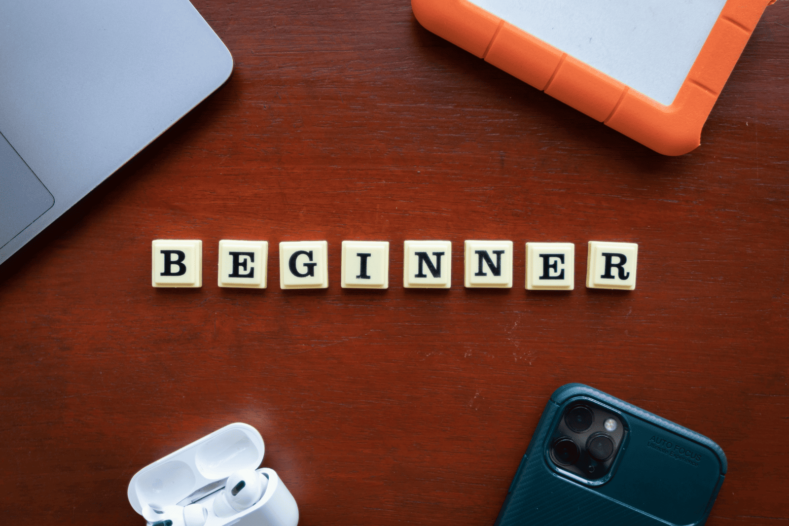 Wooden table with Scrabble tiles spelling "BEGINNER," surrounded by a laptop, phone, AirPods case, and an orange external drive, suggesting tech learning